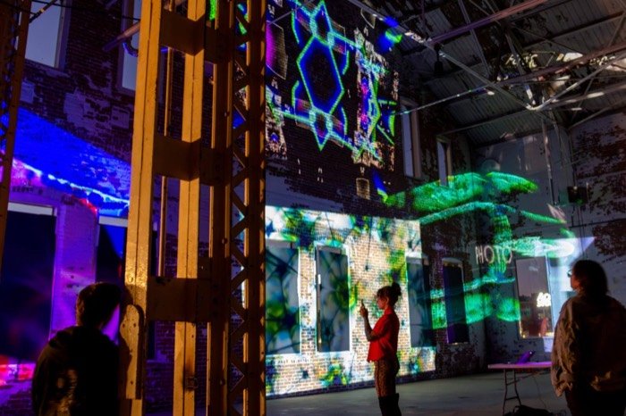 An attendee photographs overlapping green geometric and botanical projections on a brick wall at an OPTICKS Open Lab